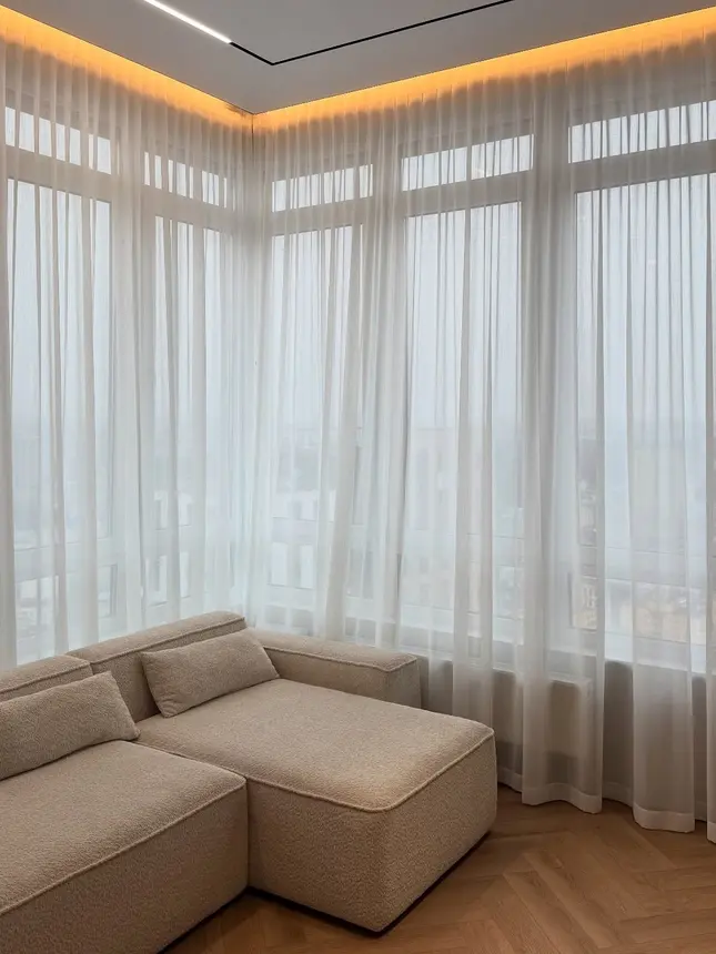 Balto tiulio užuolaidos kampiniuose panoraminiuose languose su apšviestomis lubomis ir sofa